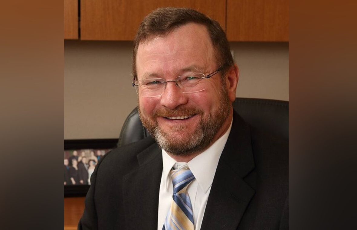 A man with brown hair and greying beard, wearing glasses and a suit and tie, smiles at the camera.